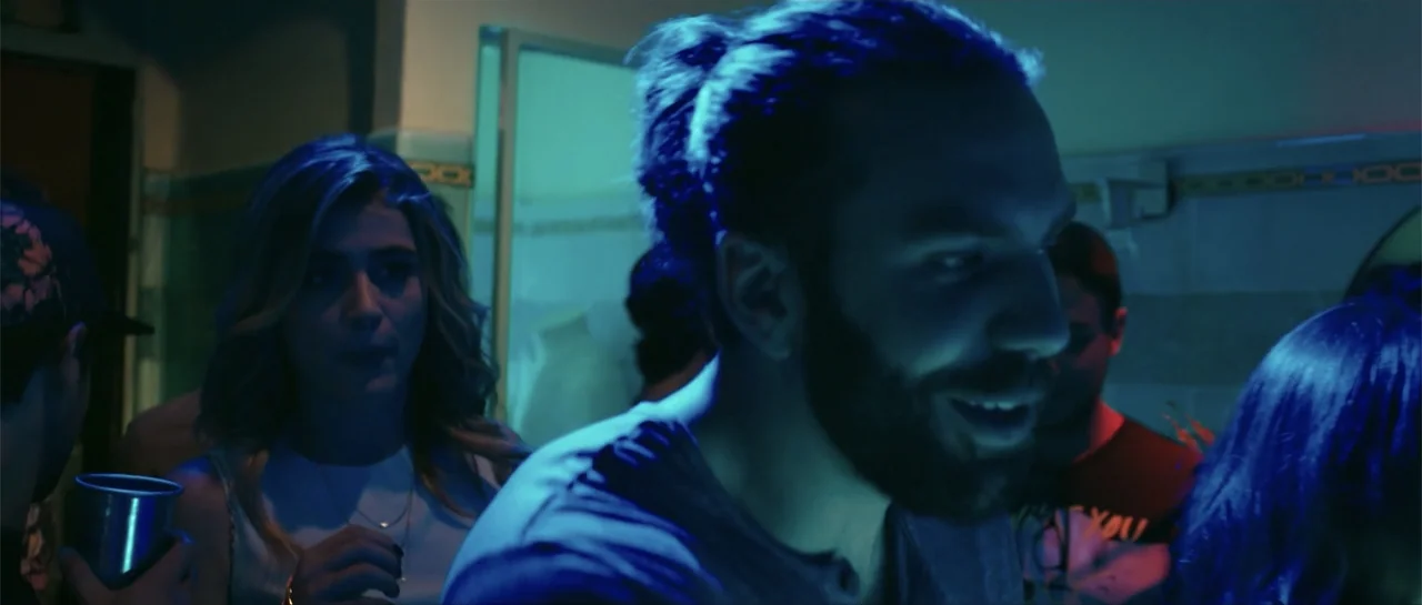 neon lighting example from Cindy La Regia: A dimly lit room filled with people during a social gathering at night. A bearded man and a blonde woman in