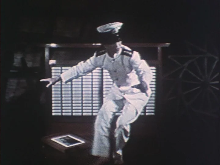 16mm film look example from The Eraser: A dimly lit room features a lattice partition in the background and a wheel. A person in a white naval uniform