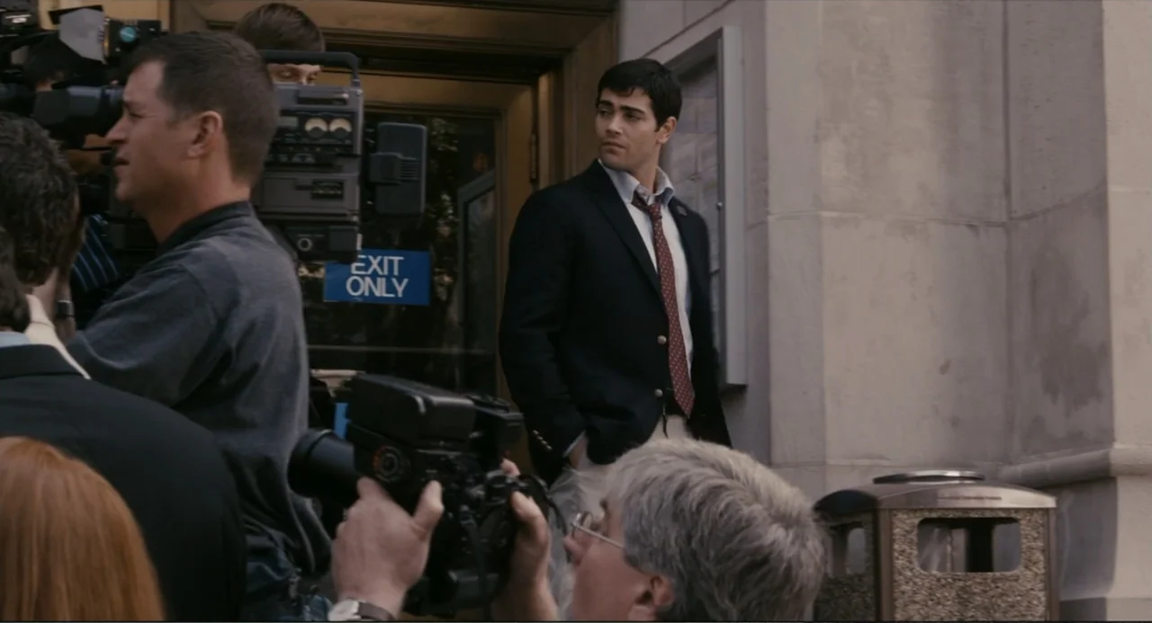 dirty single example from Beyond a Reasonable Doubt: A group of camera operators gathers outside a modern building with a concrete facade. A young man