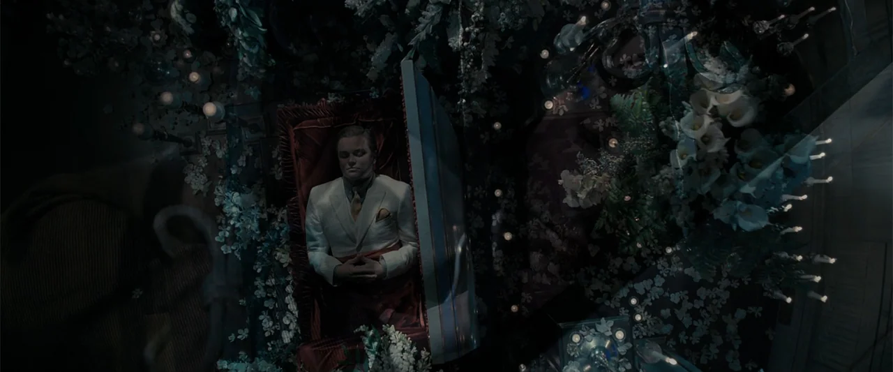 candlelight & firelight example from The Great Gatsby: A man lies inside a casket surrounded by abundant white floral sprays and candles. The camera l