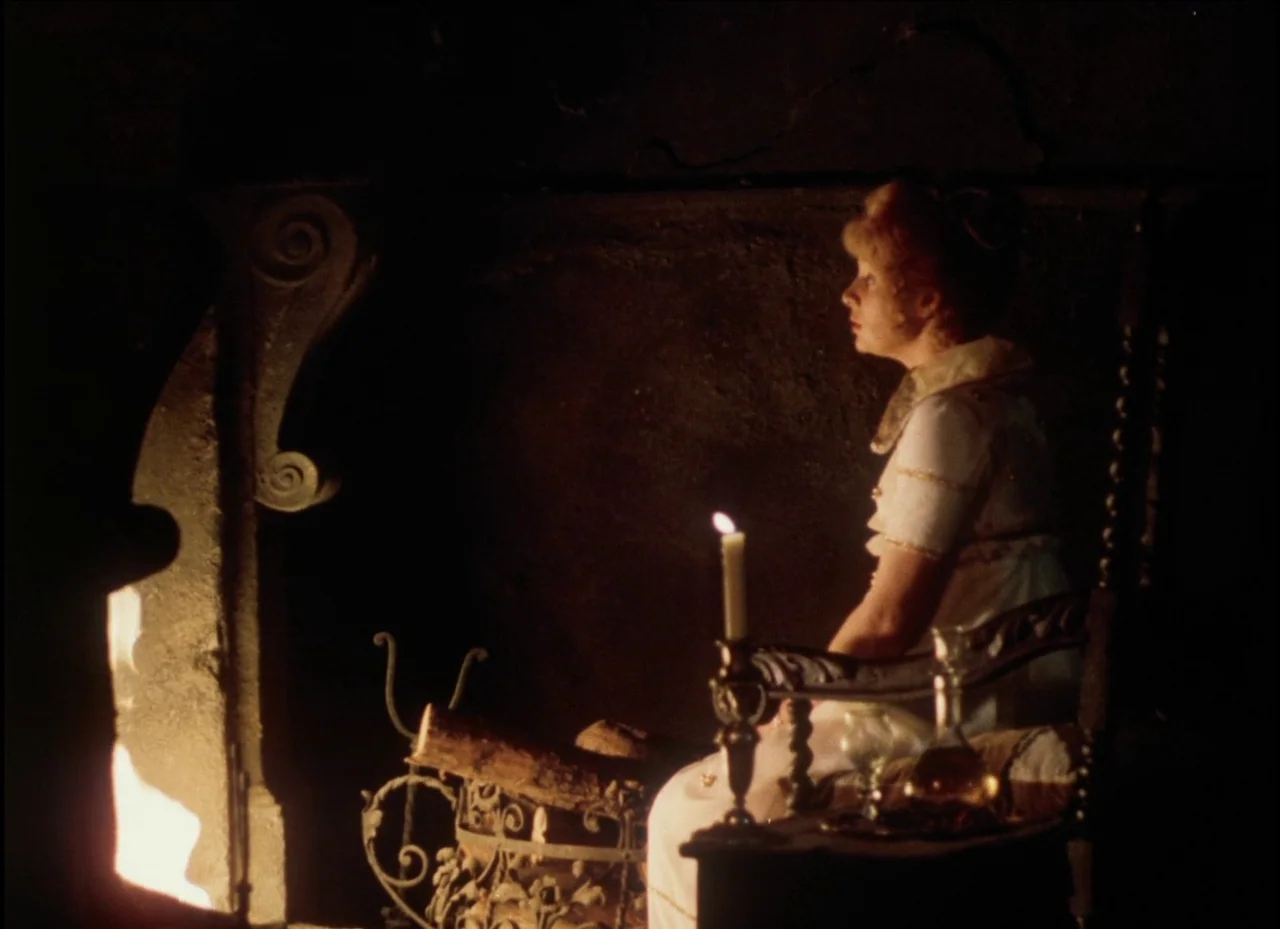 candlelight & firelight example from Beauty and the Beast: A woman in a high-waisted period dress sits in a dark, dimly lit room. She watches a firepl