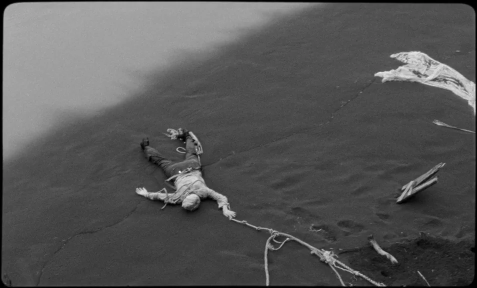overhead & bird’s-eye shots example from The Wild Boys: A dark sand dune stretches across the frame with sparse debris scattered around. A person lies