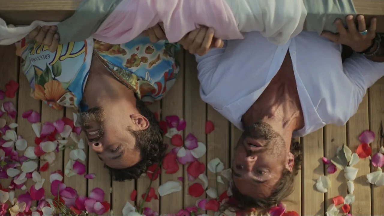 overhead & bird’s-eye shots example from Double Pregnant 2: Two men lie on a light-colored wooden deck scattered with colorful rose petals. The men lo