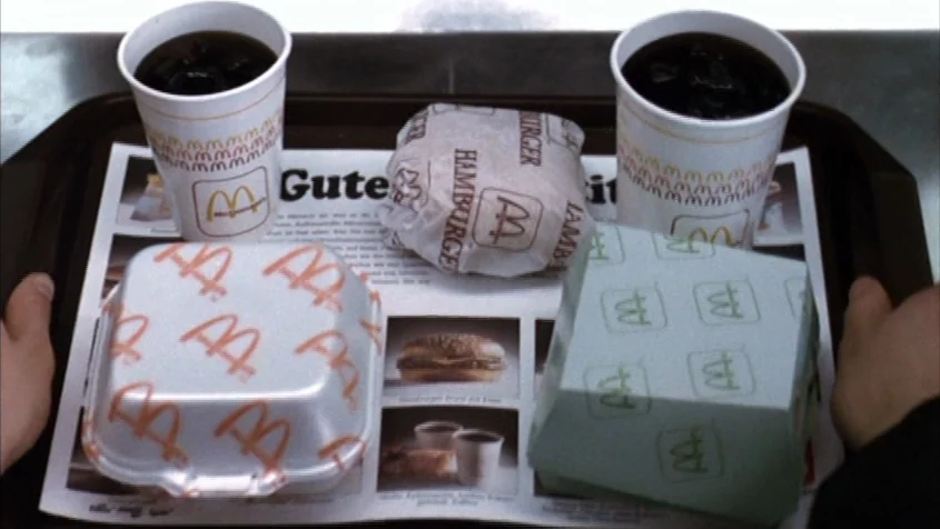 pov shot example from Benny’s Video: A plastic fast food tray holds a burger, a sandwich box, and two drinks. The items rest on a paper liner featurin