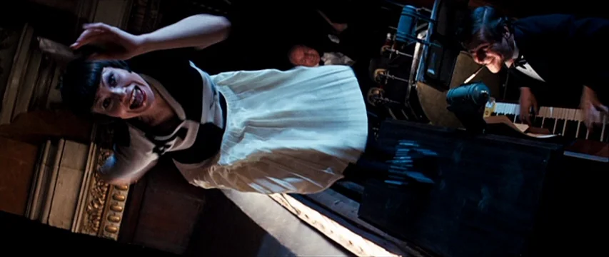 dutch angle shots example from The Boy Friend: A dark, dimly lit venue with ornate wall molding and a grand piano. A woman performs a song while looki