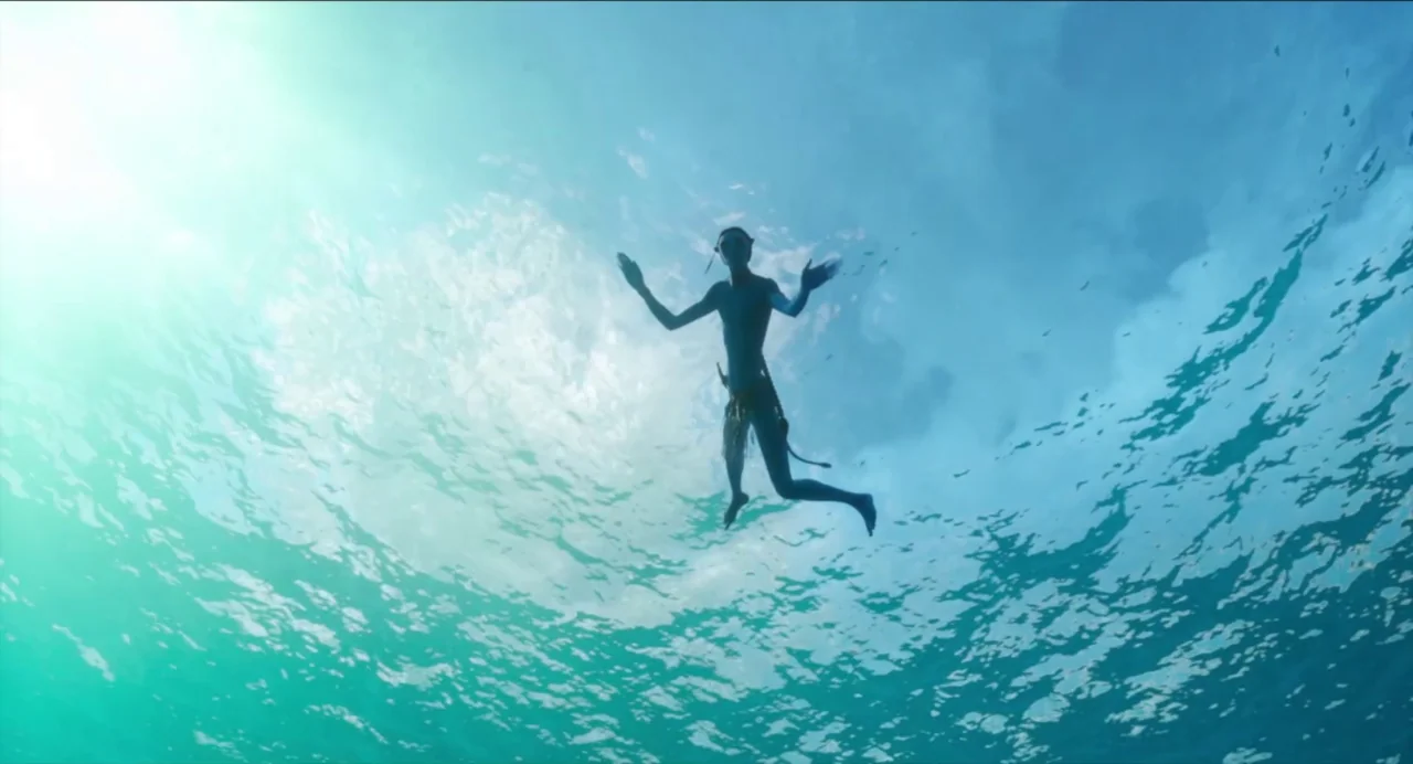 worm’s-eye shot example from Avatar: The Way of Water: A clear, sunlit ocean environment is captured from beneath the water surface. A Na'vi figure sw