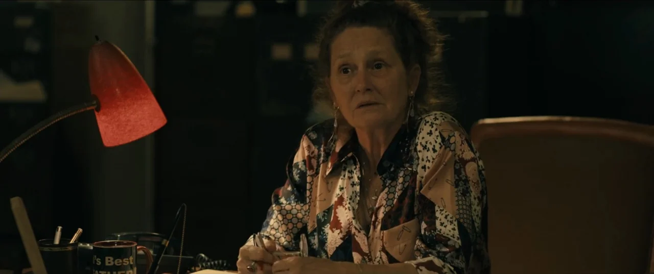 Guns Up (2025) movie still: medium — An dimly lit office space contains a desk with a red lamp and file cabinets. An older woman sits at the desk,…