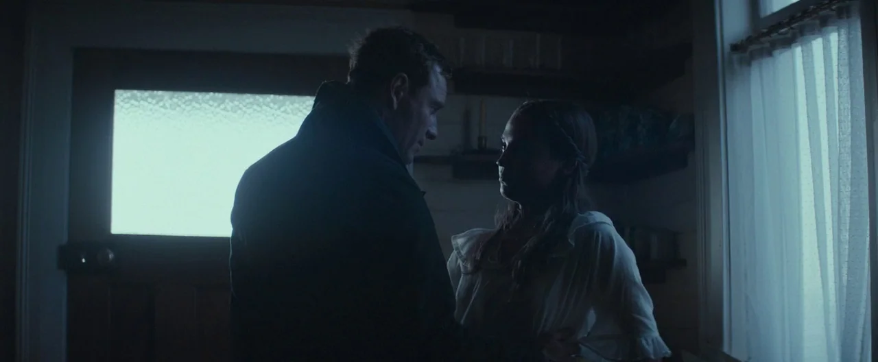 The Light Between Oceans (2016) movie still: medium — A dimly lit room in a 1920s cottage with wooden walls and window curtains. A man in a dark…