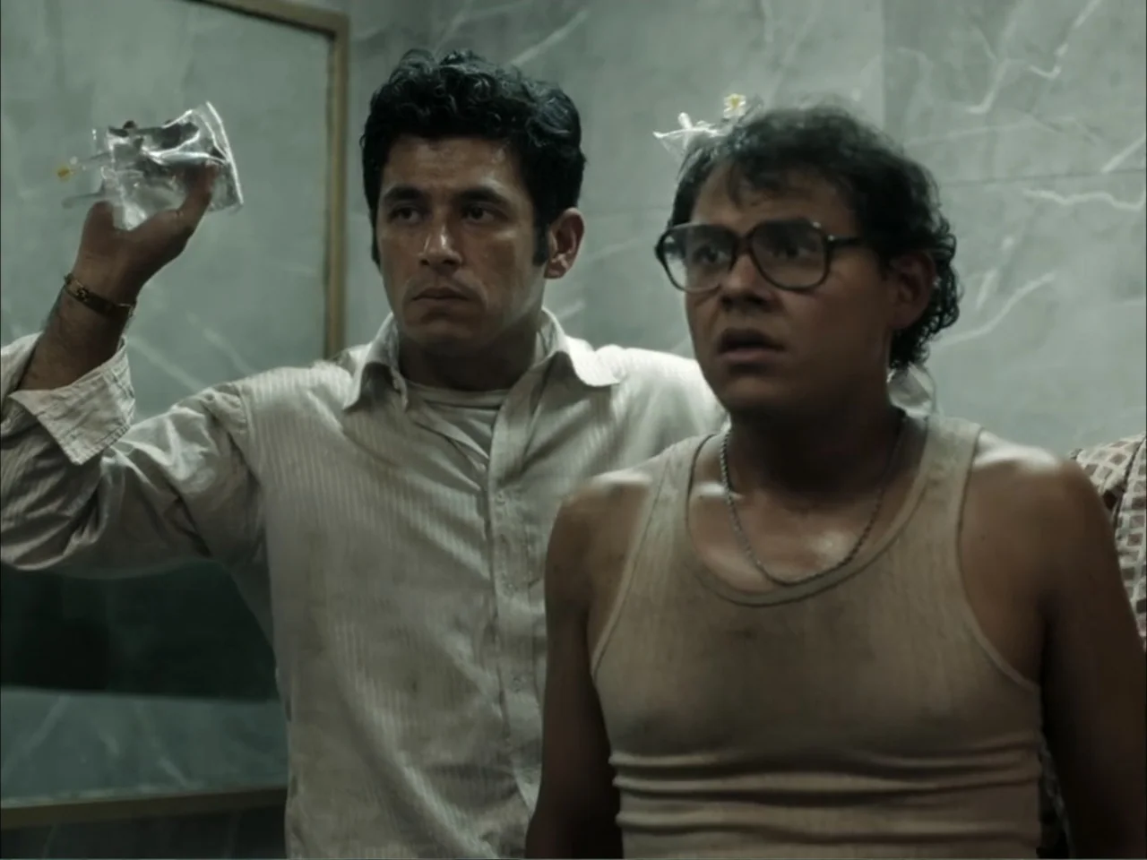 November (2025) movie still: medium — A tiled bathroom with grey marble-patterned walls contains two men standing near a mirror. The men hold small…
