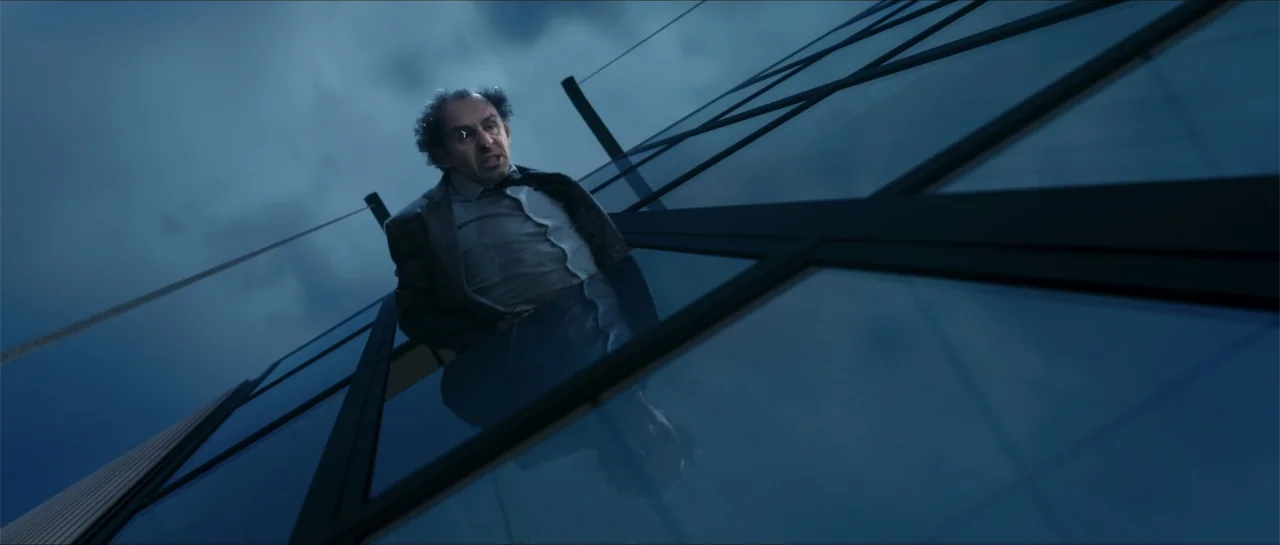 worm’s-eye shot example from Historias lamentables: A dark, modern glass skyscraper exterior is lit by cool blue ambient light. A man in a suit looks