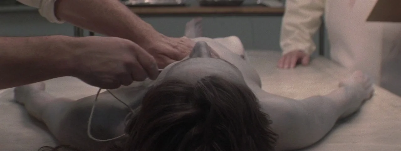 Post Mortem (2010) movie still: medium — A medical professional works on a body lying on a metal examination table. The scene is lit by flat…