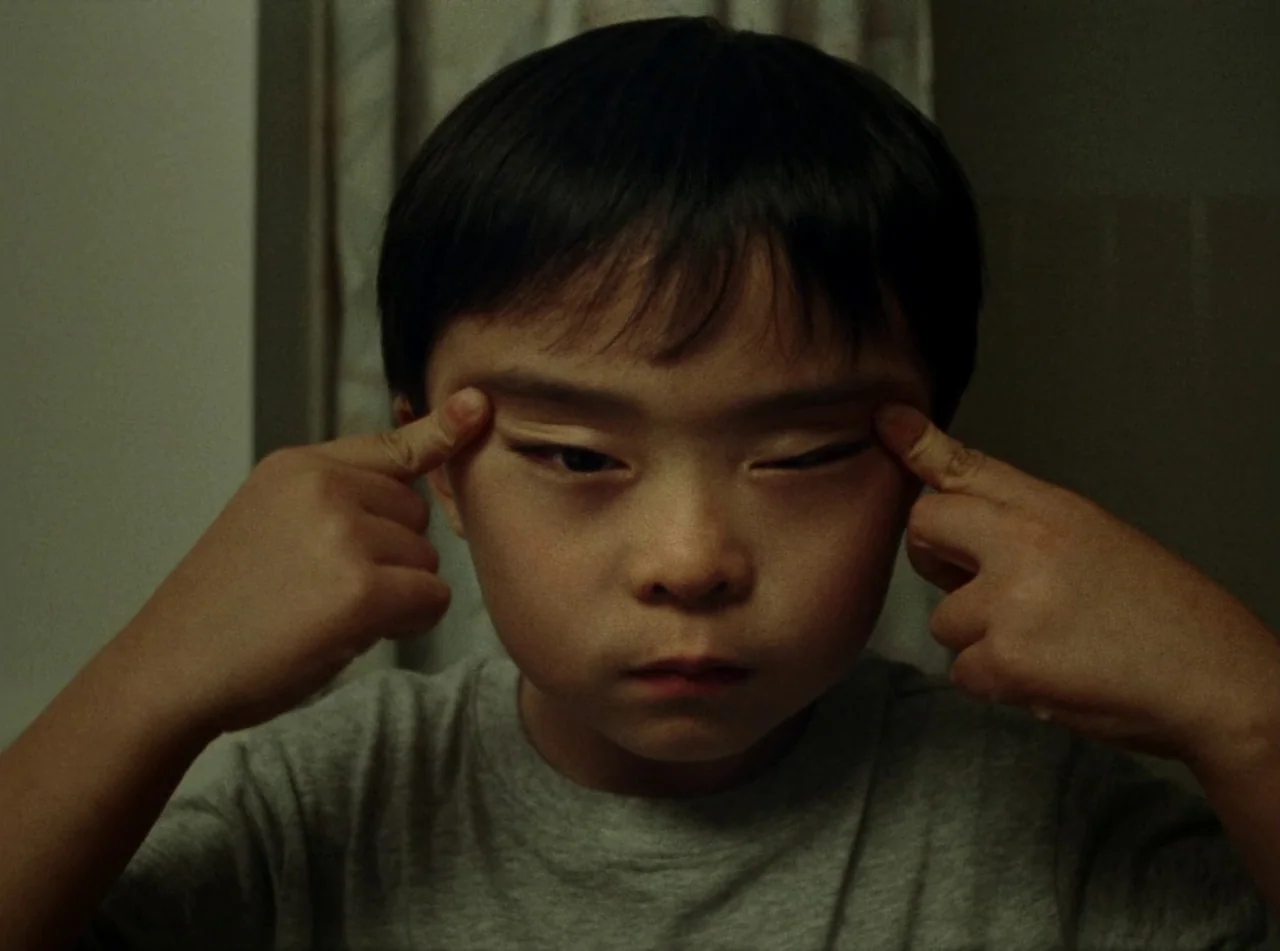Riceboy Sleeps (2022) movie still: closeup — A young boy sits indoors against a plain wall. He holds his index fingers against his upper eyelids…