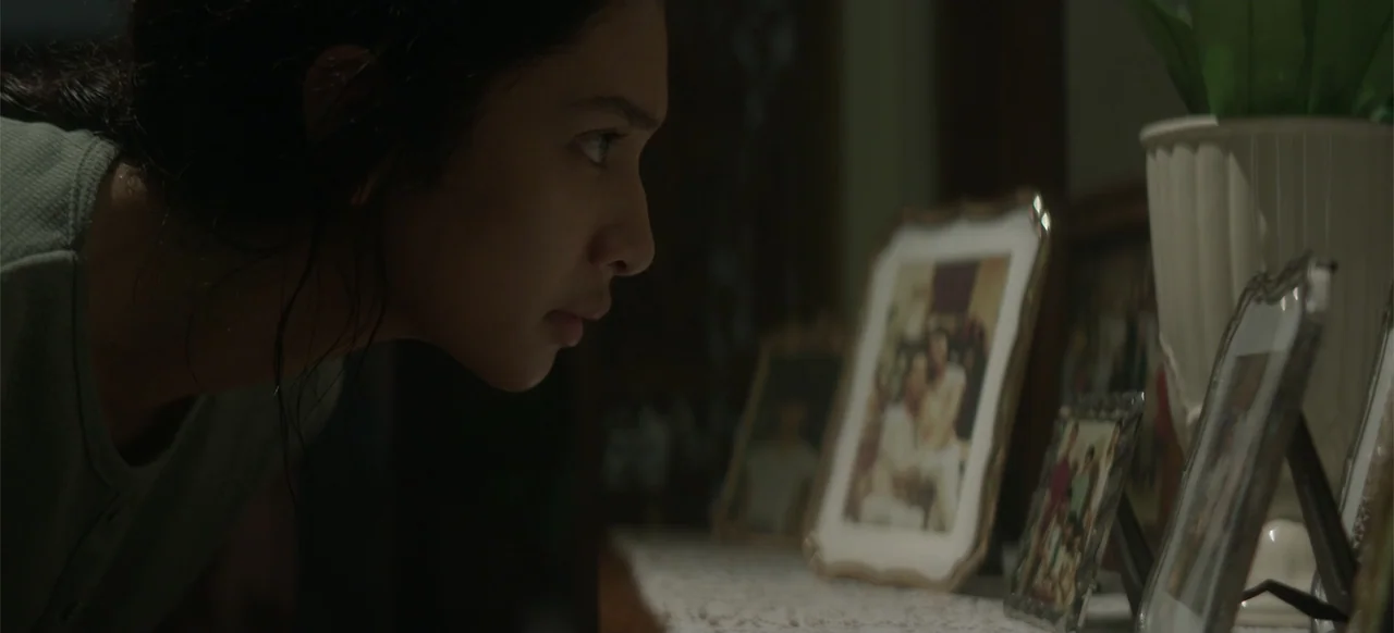 A Thousand Days (2023) movie still: closeup — A dimly lit interior room features a wooden surface covered with several framed photographs. A young…