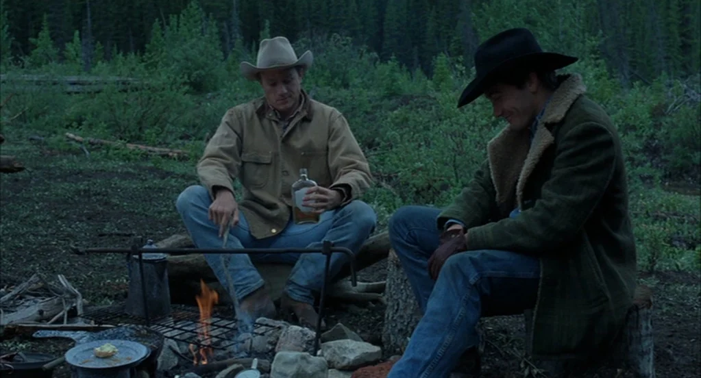 Brokeback Mountain (2005) movie still: fullbody — A dark, forested campsite with a small campfire burning in a metal grate. Two men sit on stumps…