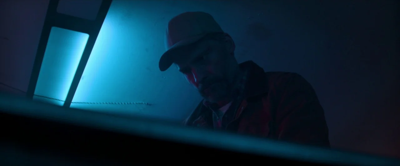 The Silencing (2020) movie still: medium — A dimly lit room is bathed in blue light from an angled neon source. A man in a flannel shirt and…