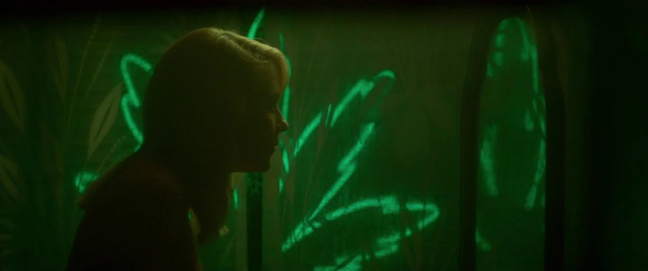 Woodshock (2017) movie still: medium — A dark interior room features walls patterned with bright green neon leaf silhouettes. A woman stands in…