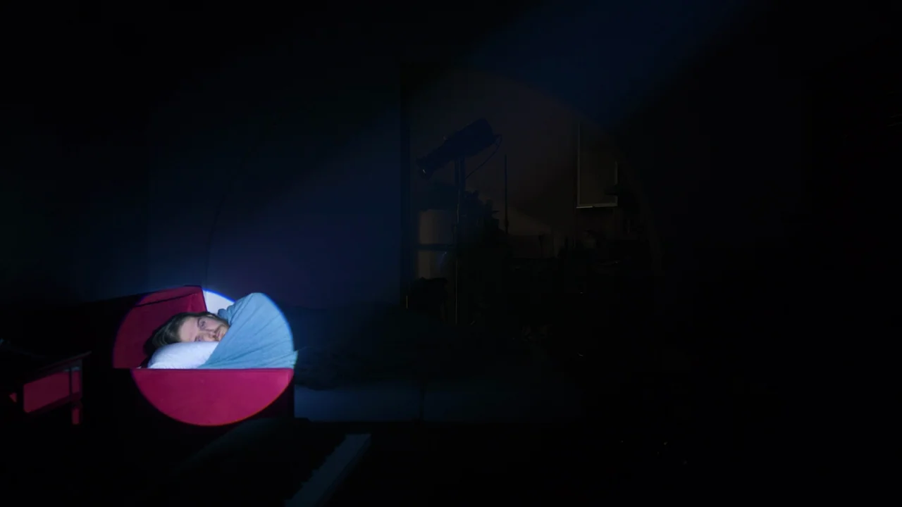 Bo Burnham: Inside (2021) movie still: wide — A dark, dimly lit room contains a person lying on a red bed frame. A tight spotlight casts a blue…