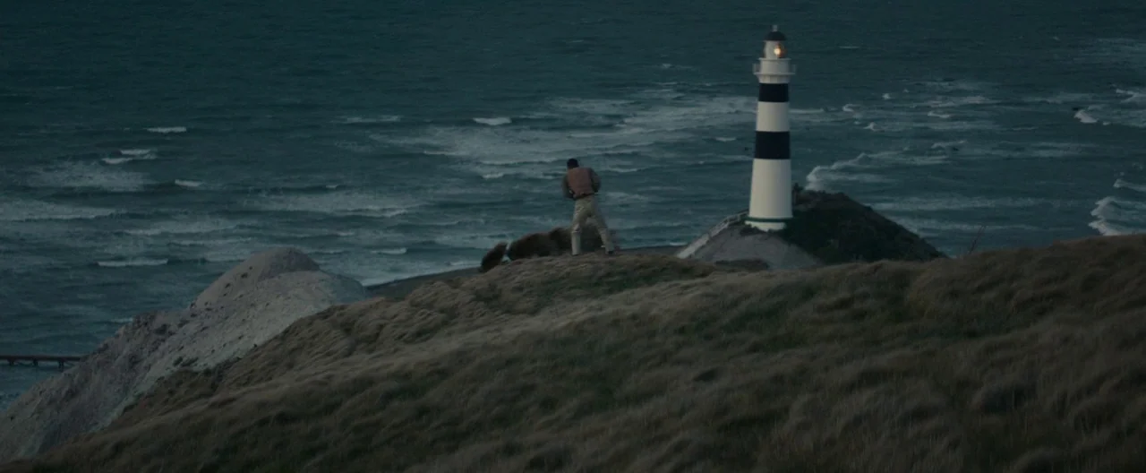 The Light Between Oceans (2016) movie still: wide — A wind-swept coastal ridge overlooks a dark ocean with a striped lighthouse nearby. A man stands…