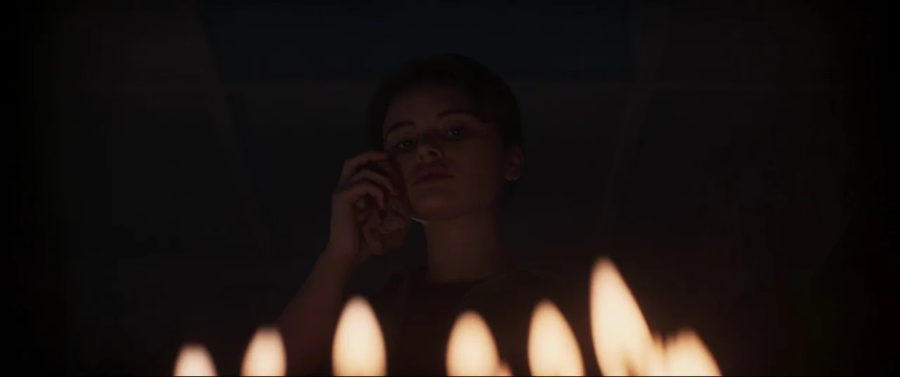 Evil Dead Rise (2023) movie still: closeup — A dark, dimly lit room is illuminated from below by a cluster of candles. A young woman holds a folded…