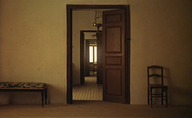 Spirit Of The Beehive (1973) movie still: wide — A series of interconnected rooms features plain beige walls and hardwood flooring. A wooden bench…