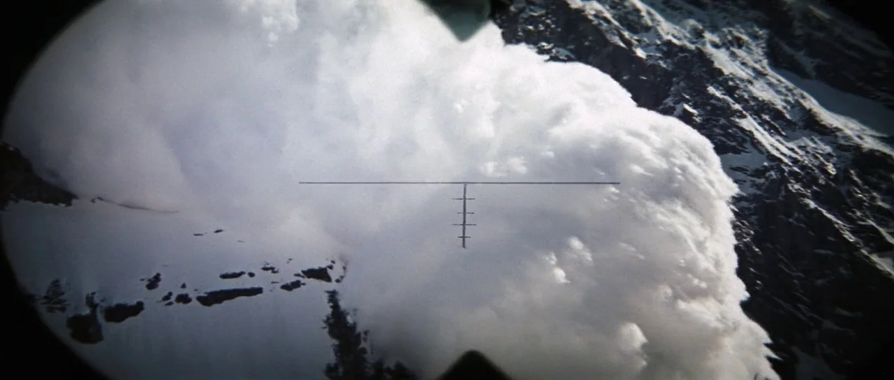 A massive white snow avalanche tumbles down a steep, rocky mountain cliff side. The view is framed through a circular reticle indicating an optical scope.