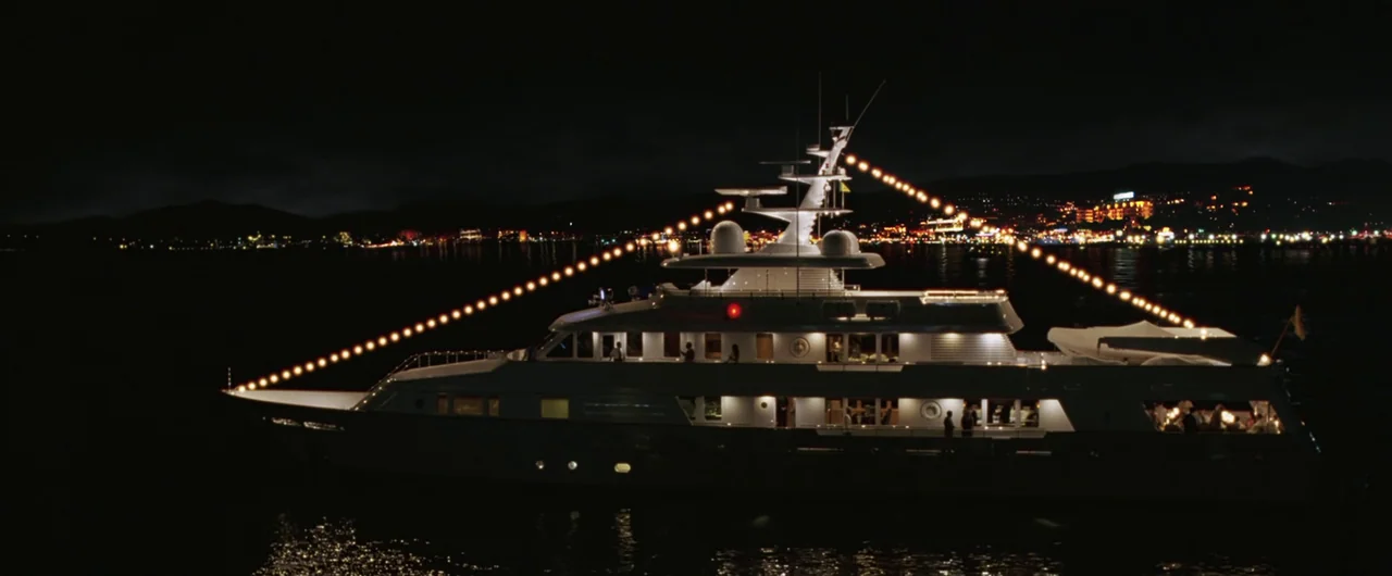 Syriana (2005) movie still: wide — A luxury motor yacht is anchored at night on dark water. Decorative golden string lights are draped from the…