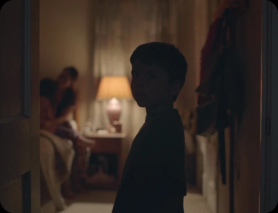 A Ghost Story (2017) movie still: medium — A dark hallway leads into a warmly lit room with a bedside lamp visible. A child stands in the foreground…