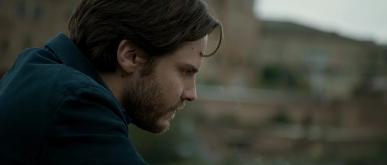 The Face of an Angel (2014) movie still: closeup — An outdoor overlook with a blurred landscape in the background under an overcast sky. A man looks…