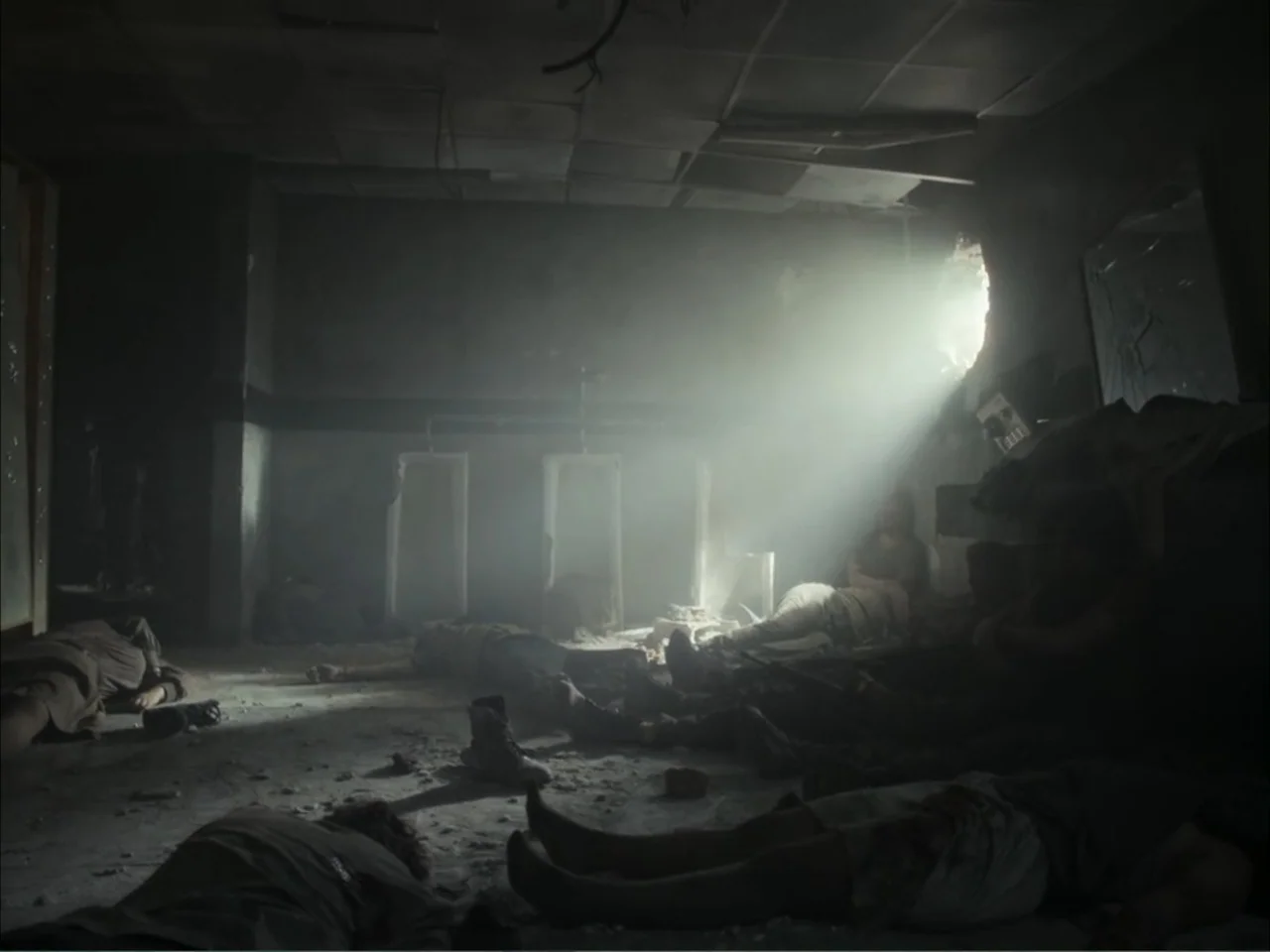 November (2025) movie still: wide — A dark, dilapidated interior room contains debris and dust-filled light streaming through a wall hole. Several…