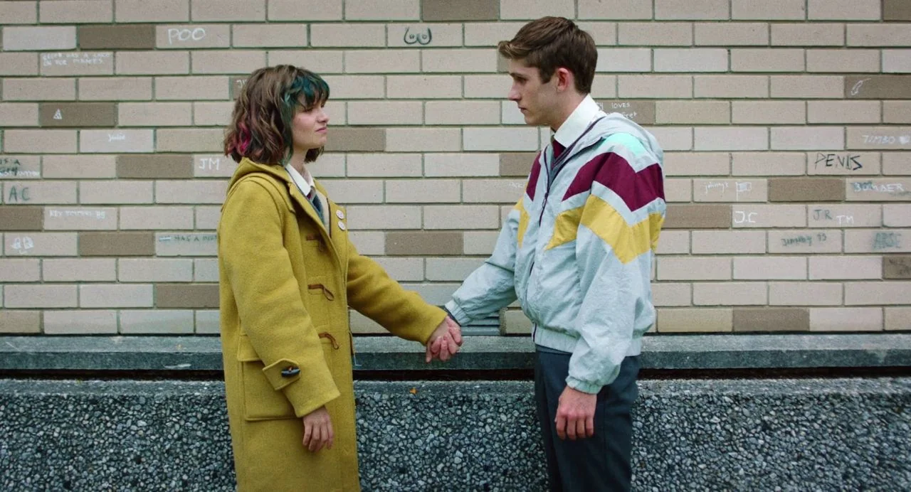 Dating Amber movie still: medium — A tan brick wall marked with casual graffiti serves as a neutral backdrop. Two teenagers in school uniforms stand…