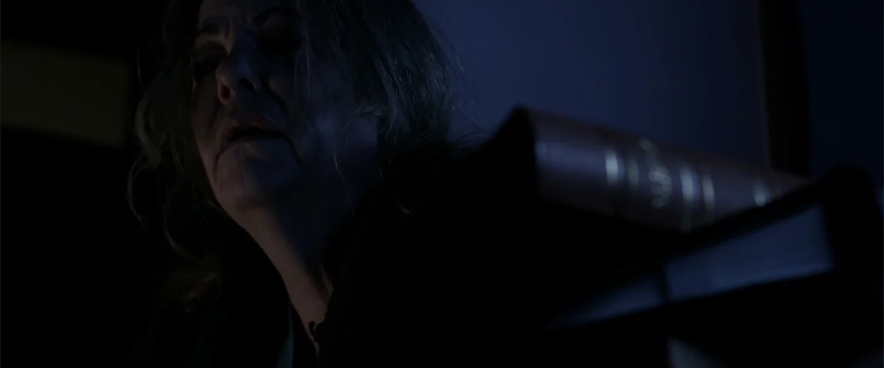 Mermaid Down (2019) movie still: closeup — A dimly lit room at night features a large closed book in the foreground. An elderly woman looks toward a…