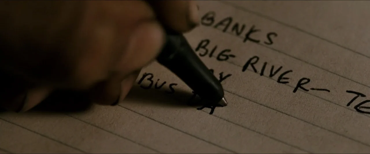 Into the Wild (2007) movie still: closeup — A close-up view shows a black ballpoint pen writing on off-white lined paper. The text lists locations…