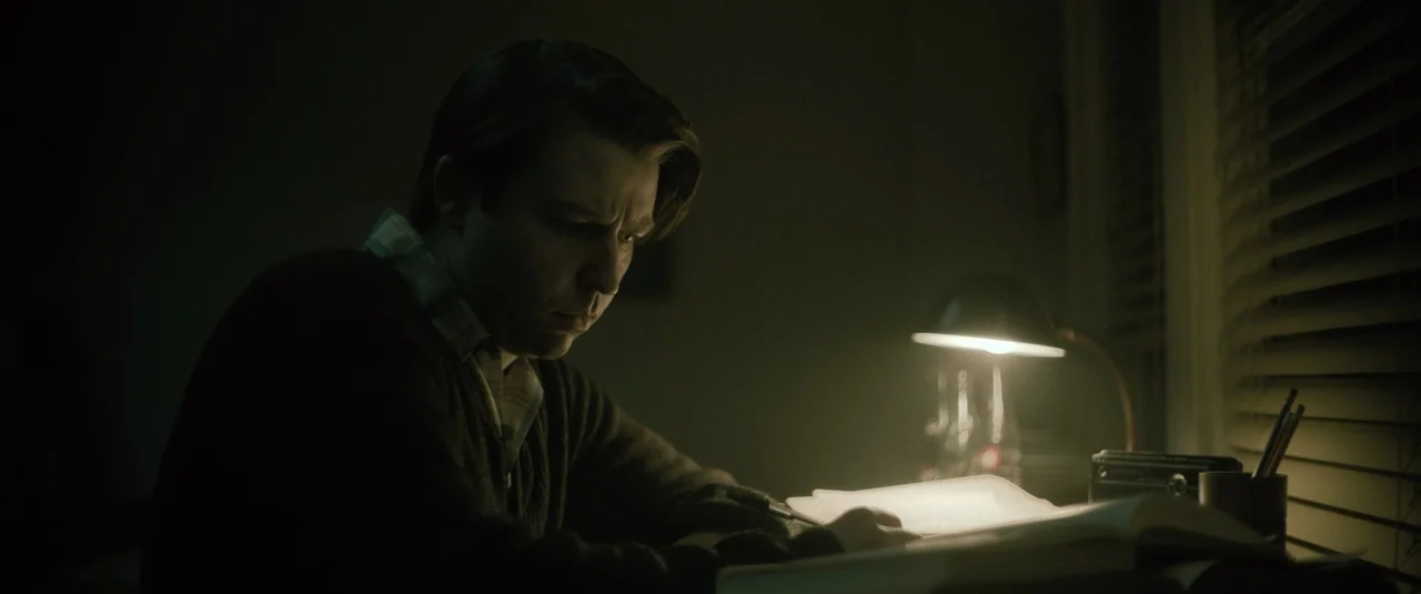 Boston Strangler (2023) movie still: medium — A dim room is illuminated only by a lamp on a cluttered desk surface. A man sits at the desk while…