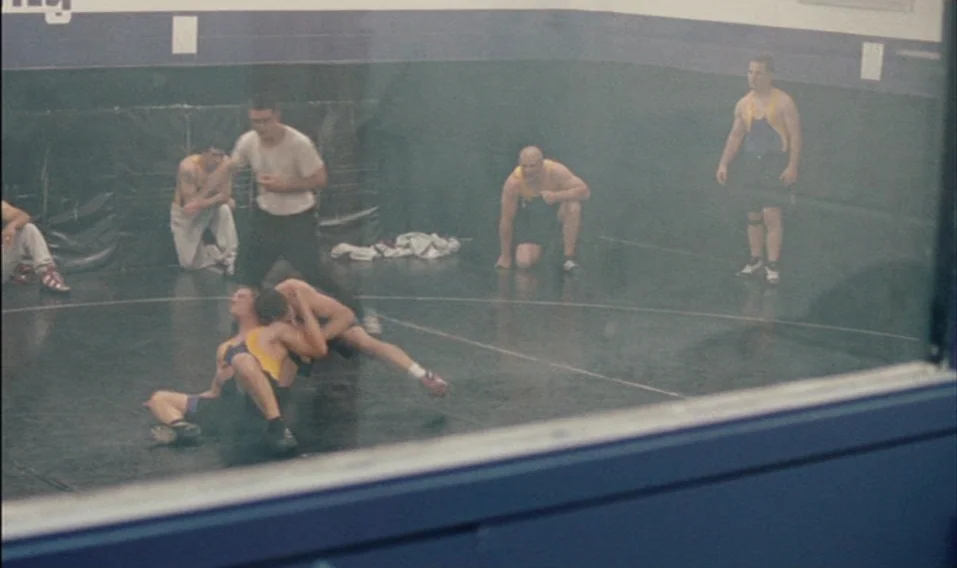 Blue Valentine (2010) movie still: wide — An indoor wrestling room with a dark mat and padded blue wall panels. Two teenagers are locked in a…