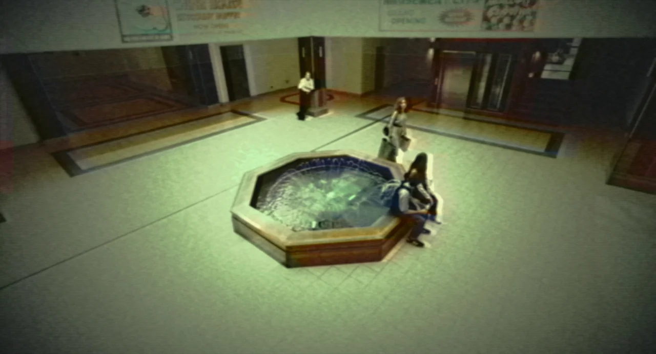 Beau Is Afraid (2023) movie still: wide — A quiet, desolate mall atrium at night featuring an octagonal central water feature. Several people occupy…