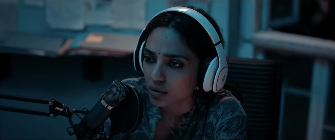 Cheekatilo (2026) movie still: medium — A dimly lit room contains recording equipment including a large microphone and headphones. A young woman…