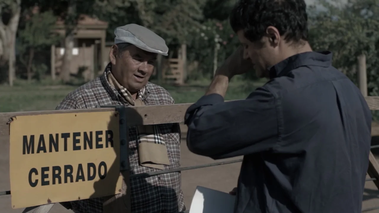 Still Life (2013) movie still: medium — An outdoor rural area with a wooden fence featuring a yellow signage board. A man with a flat cap stands…