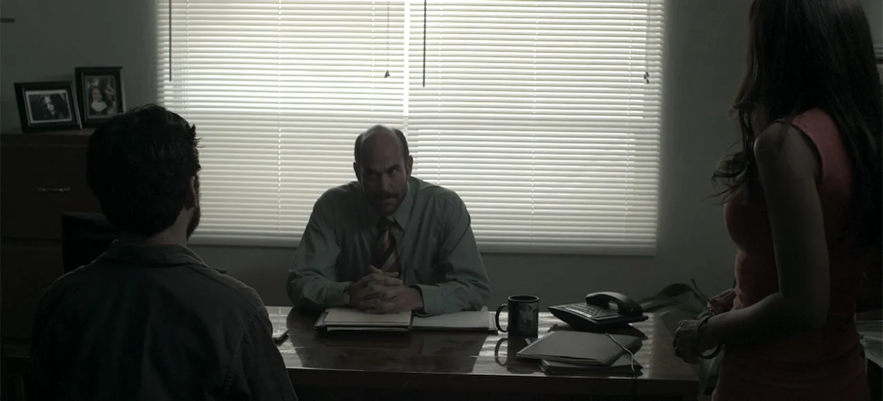 About the Deaths (2016) movie still: medium — A dark, cramped room featuring a wooden desk positioned against a window with blinds. A man in a tie…
