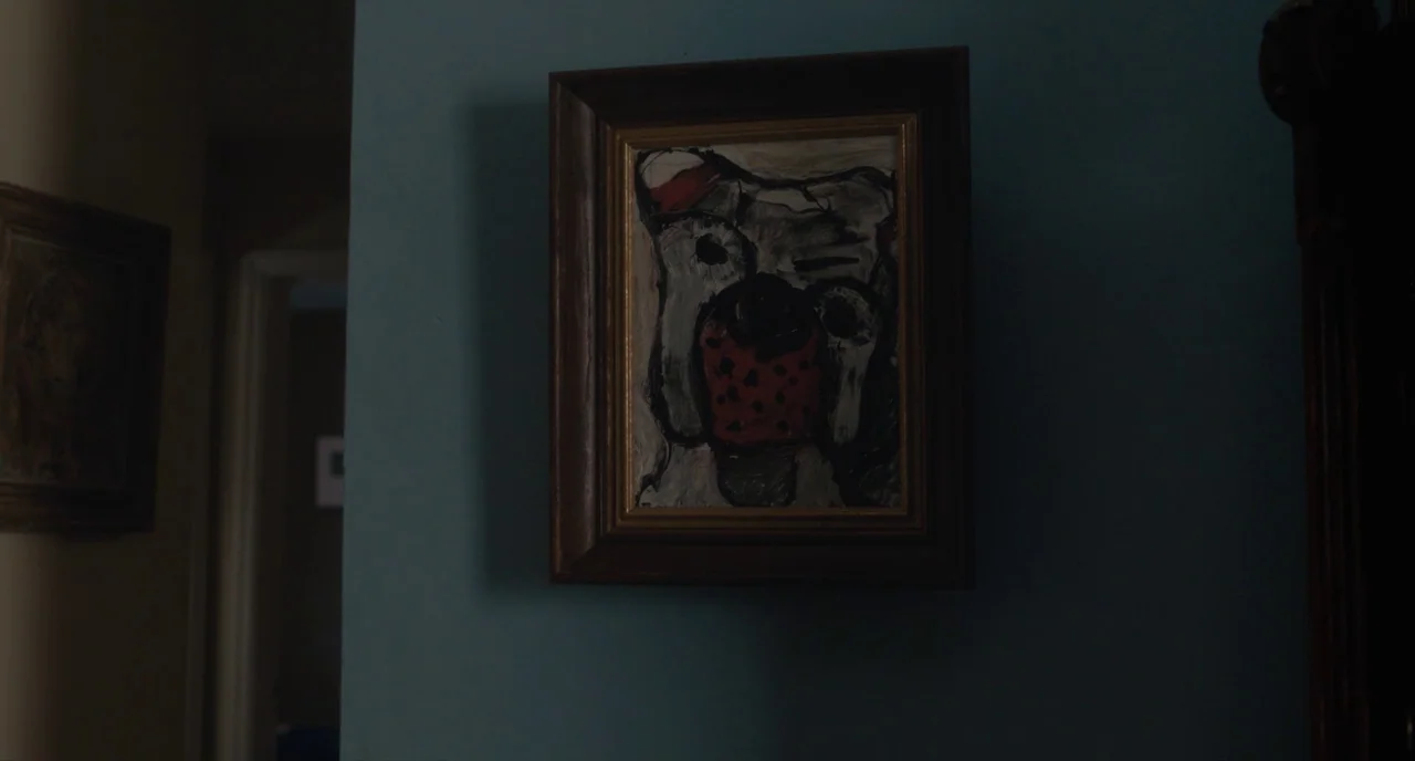 Paterson (2016) movie still: closeup — A framed abstract painting of a dog hangs on a blue-painted wall. The hallway is dimly lit with shadowed…