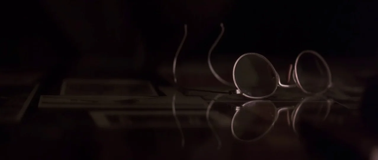 Snow Falling on Cedars (1999) movie still: closeup — A pair of thin-rimmed circular spectacles rests upon a dark, polished, reflective surface.…