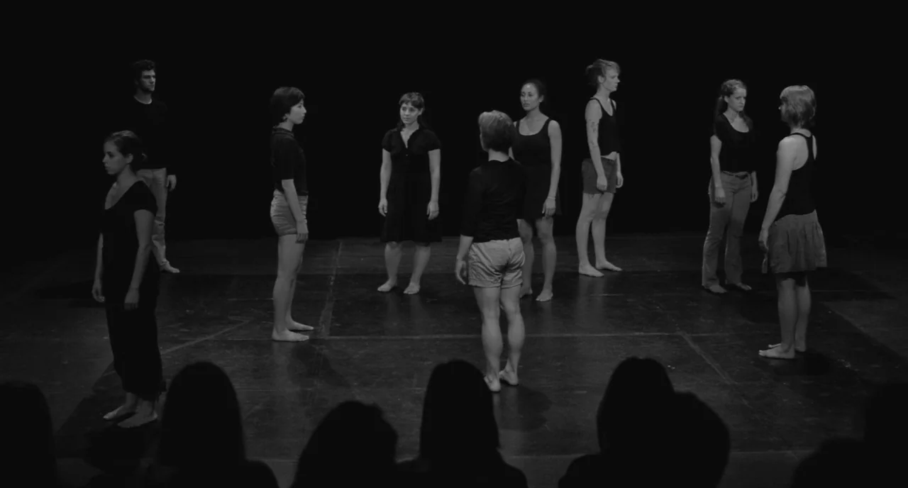 Frances Ha (2012) movie still: wide — A dark, empty stage with a black backdrop hosts eight individuals arranged in a circle. They are all barefoot…