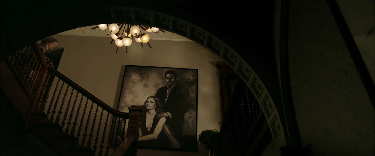 K.G.F: Chapter 2 (2022) movie still: wide — A dark staircase landing in a house features a large portrait on the wall. A woman stands on the upper…