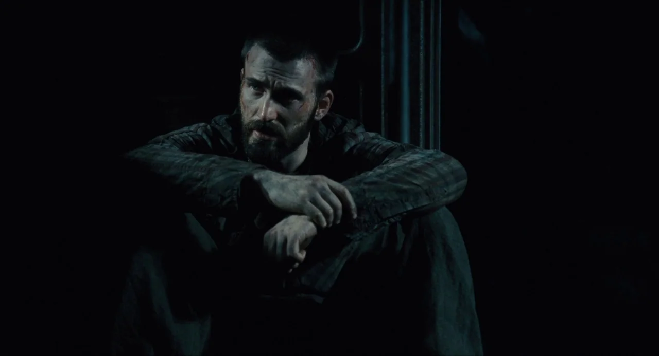 Snowpiercer (2013) movie still: medium — A dark, minimally lit space with a structural metal pillar visible in the background. A man with a bruised…