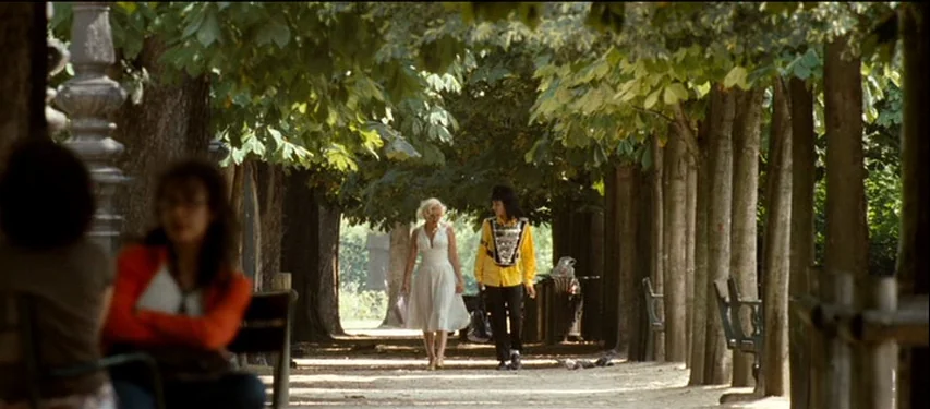 Mister Lonely (2007) movie still: wide — A gravel pathway cuts through a dense line of trees in a public park. A woman in a white dress walks…