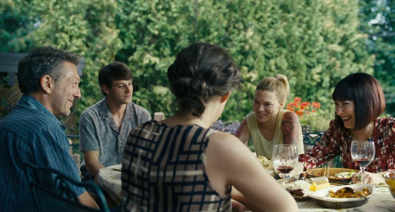 It’s Only The End Of The World (2016) movie still: medium — A group sits around a round table on a leafy outdoor patio. They are sharing a meal and…