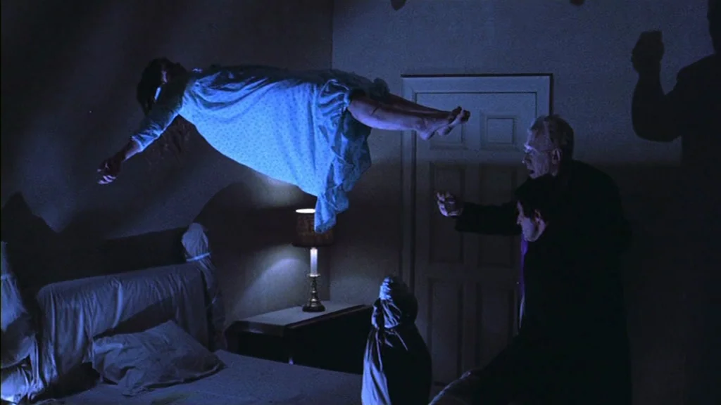 The Exorcist (1973) movie still: wide — A dimly lit room features a bed and a small table with a lamp. A girl floats in mid-air while two men watch…