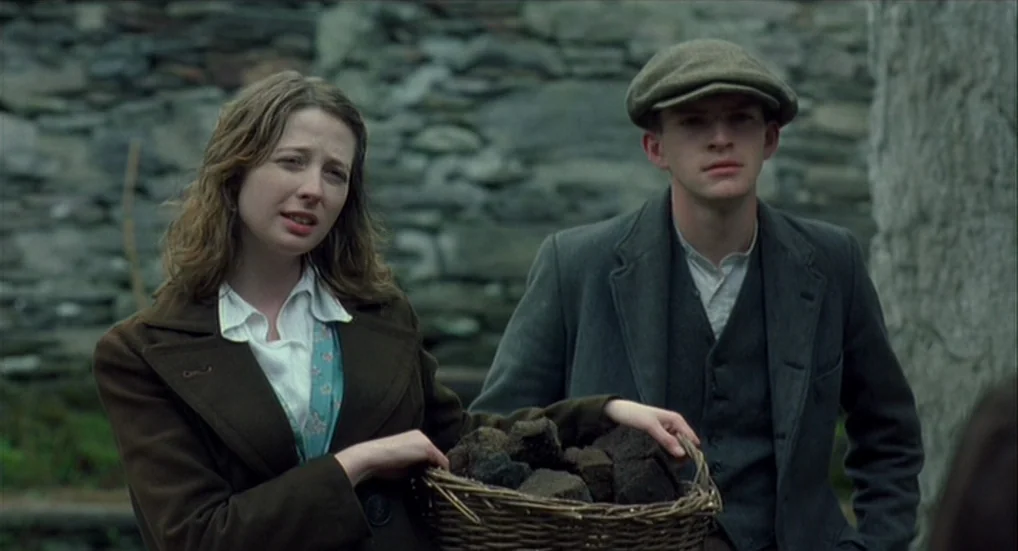 The Wind That Shakes the Barley (2006) movie still: medium — Two young people stand in front of a rustic, stacked dry-stone wall. The woman holds a…