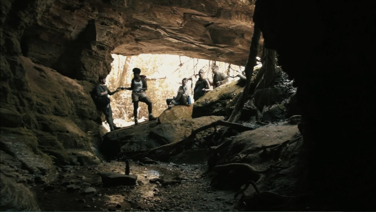 The Grotto (2022) movie still: wide — A group of five hikers congregates inside a large rocky cave opening with textured walls. They stand on uneven…