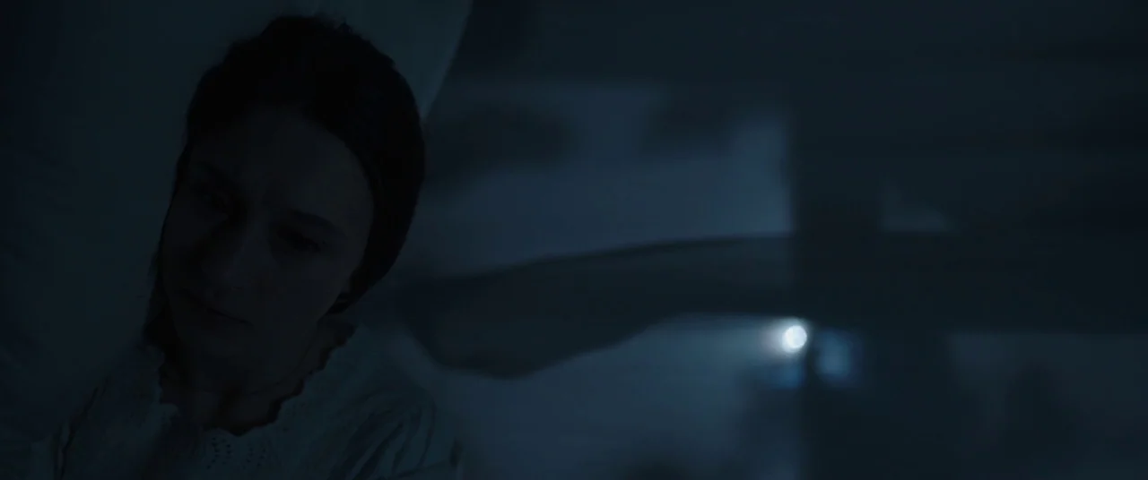 The Nun (2018) movie still: closeup — A dimly lit room at night features a woman lying in a bed. She gazes out into the darkness near a window…