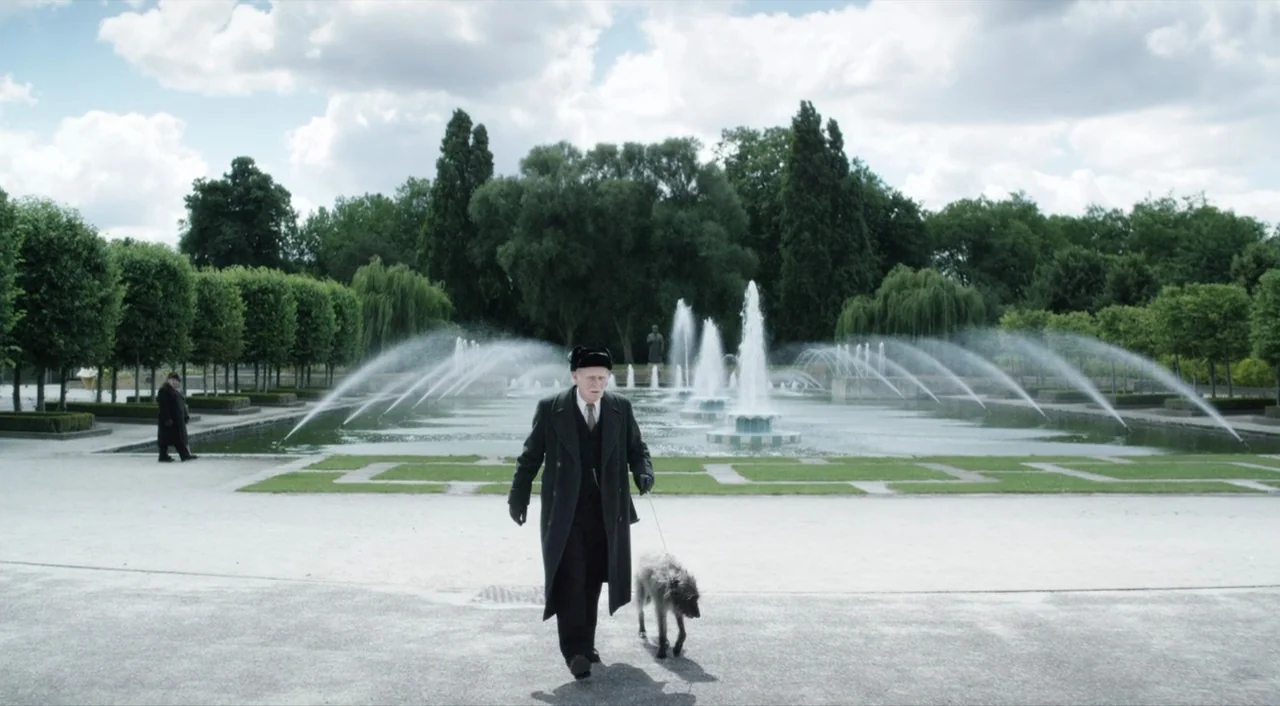 The Death of Stalin (2017) movie still: fullbody — A man in a winter coat walks with his dog near a park fountain. Behind him a smaller figure…